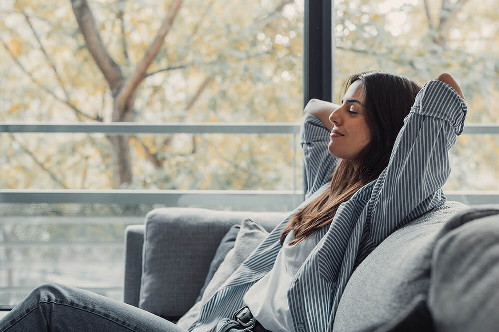 Entspannte Frau sitzt auf einer Couch, Augen geschlossen, Arme hinter dem Kopf, leichtes Lächeln im Gesicht