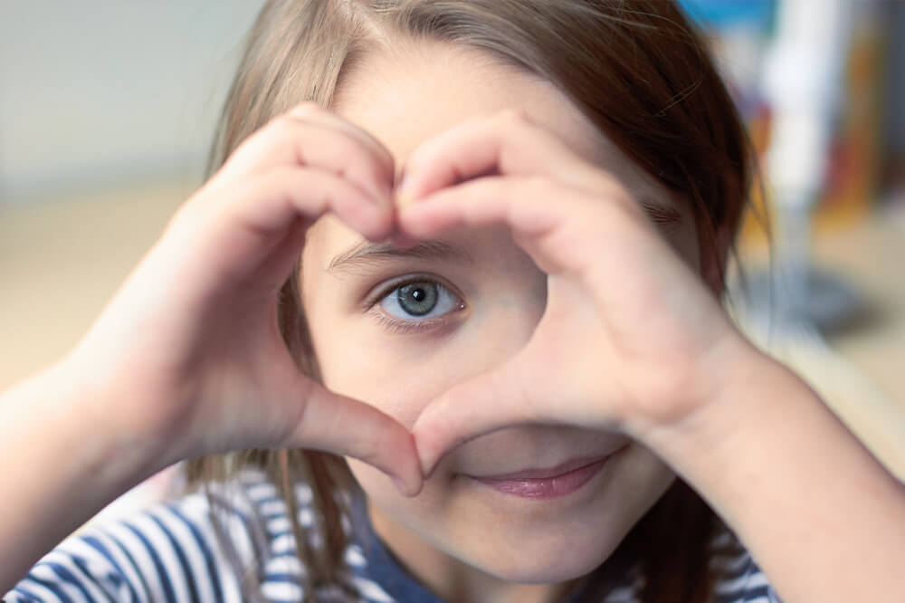 Junger Junge hält die Hände zu einer Herzform vor dem Gesicht und schaut mit einem Auge hindurch